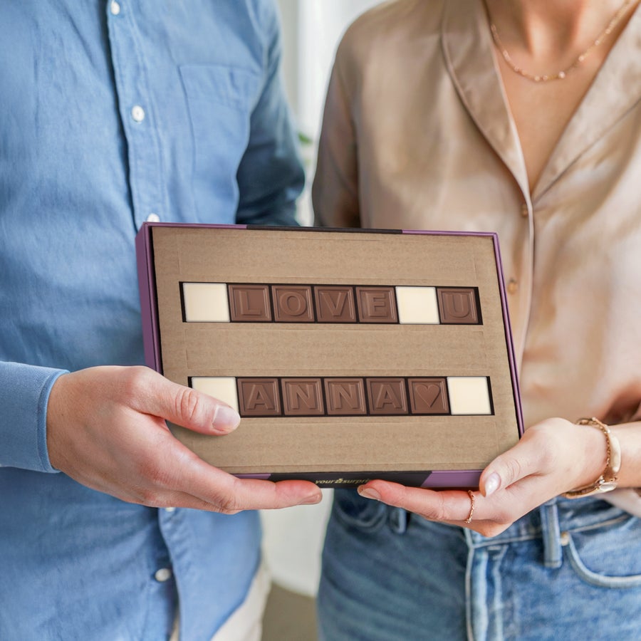 A couple holding an open gift box with a personalized chocolate telegram that spells LOVE U ANNA in milk chocolate letters.