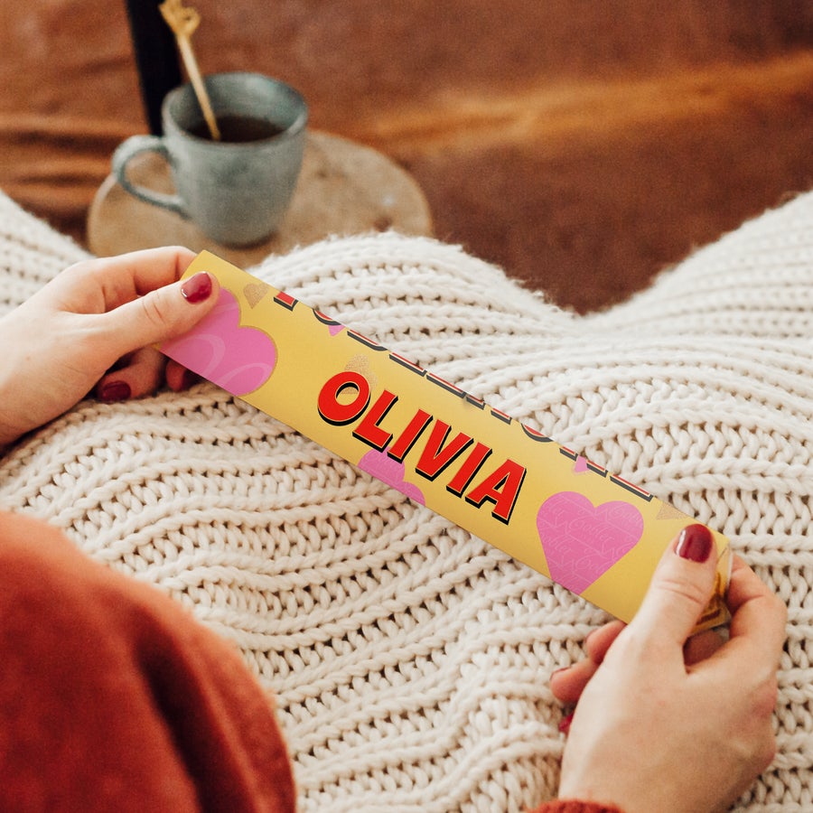 Hands holding a personalized Toblerone bar with the name Olivia printed on a yellow wrapper, featuring pink heart designs